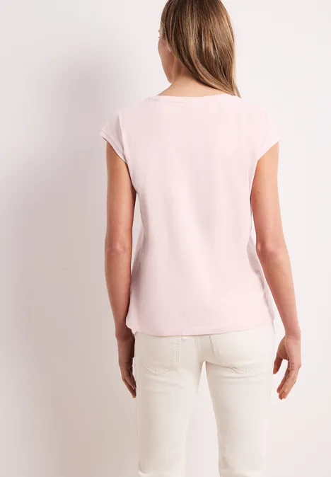 Frau von hinten in hellrosa T-Shirt und cremefarbenen Hosen vor hellem Hintergrund.